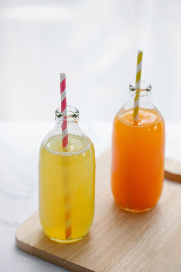 Fresh orange and yellow juice in glass bottles with straws on a wooden tray.